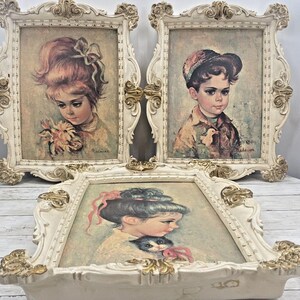 May include: Three vintage framed portraits. Two are vertical, one horizontal. The frames are ornate, cream-coloured with gold accents. The portraits depict children with soft, muted colours. One girl holds flowers, another a cat, and the third a boy in a cap.