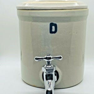 Könnte beinhalten: Ein beigefarbener Wasserbehälter aus Steinzeug mit Deckel und verchromtem Wasserhahn. Der Kühler hat ein dunkelblaues "D" auf der Vorderseite. Der Deckel hat einen kleinen Knopf.
