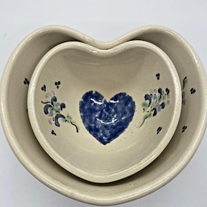 May include: A set of two heart-shaped ceramic bowls. The smaller bowl is nested inside the larger bowl. Both bowls are decorated with a blue heart and floral designs in shades of blue, green, and pink.
