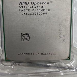 Op de afbeelding: Een vierkante AMD Opteron processor met een zilverkleurige metalen heatspreader met de tekst AMD Opteron, OSA254FAA5BL, CABYE 0526WPMW, Y936283G50066 en ASSEMBLED IN MALAYSIA. De processor heeft een groene printplaatbasis en een datamatrixcode.