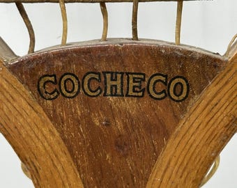 Könnte beinhalten: Nahaufnahme eines Vintage-Tennisschlägers aus Holz. Der Schläger hat einen braunen Holzrahmen mit dem Wort "COCECO" in Schwarz auf dem Griff. Die Saiten sind hellbraun, und das Design ist klassisch und nostalgisch.