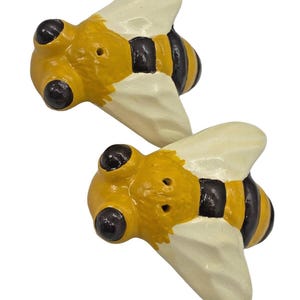 Pode incluir: Dois objetos decorativos em forma de abelha. Cada abelha tem um corpo amarelo com listras pretas, olhos grandes pretos e asas esbranquiçadas. As abelhas são pintadas com um acabamento texturizado, dando-lhes uma aparência artesanal.