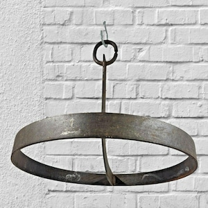 May include: A circular metal ring with a dark, weathered finish, featuring a vertical support bar that connects to a top hanging loop. The object is constructed from flat, wide metal bands and displays a textured, aged appearance throughout its surface.
