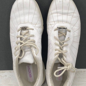 May include: A pair of white leather sneakers with a lace-up closure. The shoes have a white sole and a small metal logo on the side. The inside of the shoe has a pink label that says "Body".