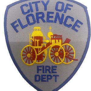 May include: A shield-shaped patch with the words "CITY OF FLORENCE FIRE DEPT" in blue lettering. The patch features an embroidered image of a vintage fire engine in red and yellow.