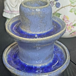 May include: A blue and white ceramic candle holder with a wide base and a smaller top opening. The holder has a glazed finish and a blue rim.