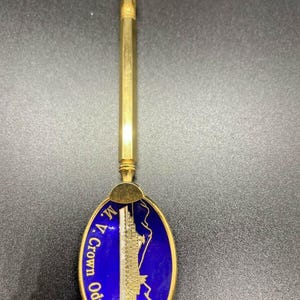 Pode incluir: Colher dourada com um design de esmalte azul que apresenta um navio de cruzeiro e o texto "M.V. Crown Odyssey".