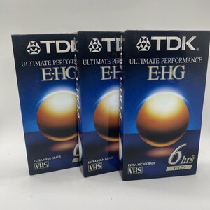 Pode incluir: Três fitas de vídeo VHS TDK azuis e prateadas com o texto "ULTIMATE PERFORMANCE EHG" e "EXTRA HIGH GRADE VHS". A fita à direita tem o texto "6 hrs T-120".