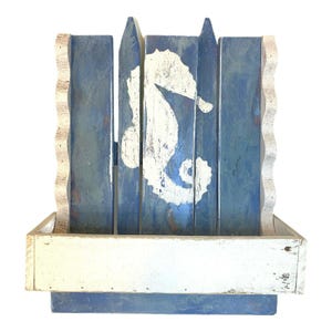 May include: A distressed, wooden wall shelf with a coastal theme. The shelf features a blue and white seahorse design on a blue background. The shelf has a white, rectangular base and wavy white trim.
