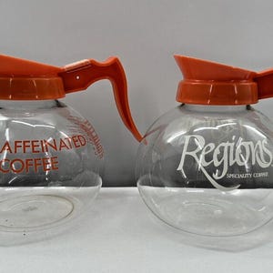 May include: Two glass coffee carafes with orange lids and handles. One carafe is labeled "Decaffeinated Coffee" and the other is labeled "Regions Specialty Coffee".