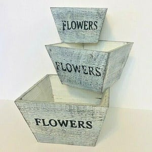 May include: A set of three nested square wooden planters with a distressed light gray and white finish. Each planter features the word FLOWERS printed in black capital letters on the front side, showcasing a weathered texture throughout.