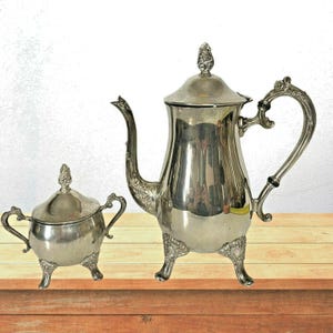 May include: A silver tea set comprising a teapot and sugar bowl, presented on a wooden surface. The teapot features an elaborate handle and spout, while both items have decorative feet and lids. The set is likely vintage or antique, with a reflective surface.