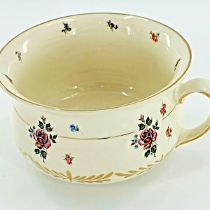 Puede incluir: Orinal de cerámica color crema con asa y diseño floral. El orinal tiene un borde dorado y detalles de hojas doradas. El patrón floral incluye flores rojas, rosas y azules con hojas verdes.