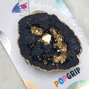 Op de afbeelding: Zwarte en gouden PopSocket PopGrip met een geode-ontwerp. De PopGrip is bevestigd aan een witte kaart met de woorden "PopSockets" en "dynamic sparkle" erop gedrukt.