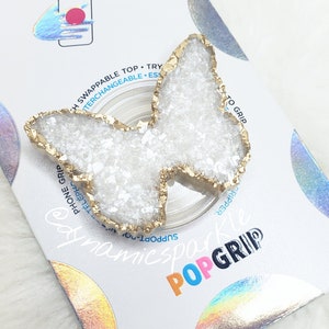 Peut inclure: Une poignée de téléphone en forme de papillon blanche avec une finition pailletée dorée. La poignée est fixée à un anneau de téléphone transparent. Le texte "POPGRIP" est imprimé sur l'emballage.