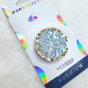 Puede incluir: Un agarre para teléfono de purpurina dorada y blanca con el texto "POPGRIP PREMIUM" en el embalaje. El agarre está diseñado para ser usado con una funda de teléfono.