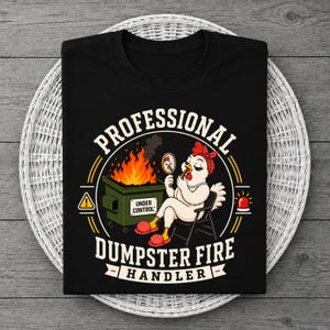 May include: Black t-shirt with a humorous graphic. The design features a cartoon chicken applying makeup while sitting next to a dumpster fire. Text reads "PROFESSIONAL DUMPSTER FIRE HANDLER."