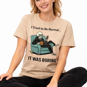 May include: Beige t-shirt with the text "I Tried to Be Normal... IT WAS BORING." The graphic shows a skeleton in a hoodie sitting in a teal armchair, holding a phone. The skeleton has a coffee mug that says "ME".