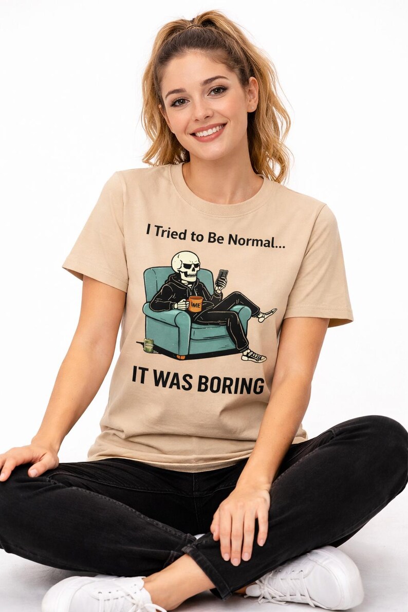 May include: Beige t-shirt with the text "I Tried to Be Normal... IT WAS BORING." The graphic shows a skeleton in a hoodie sitting in a teal armchair, holding a phone. The skeleton has a coffee mug that says "ME".
