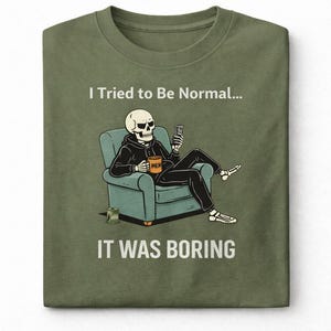 May include: Olive green t-shirt featuring a skeleton in a hoodie, seated in a teal armchair, holding a phone and a mug that says "MEM". The text "I Tried to Be Normal... IT WAS BORING" is printed above and below the illustration.