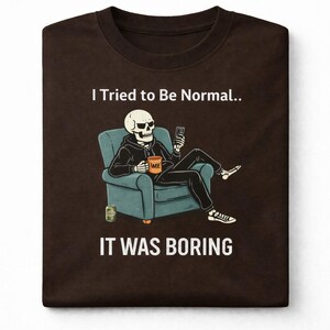 May include: Brown t-shirt featuring a skeleton in a hoodie, seated in a teal armchair, holding a phone. The skeleton has a coffee mug that says "ME". The text on the shirt reads "I Tried to Be Normal.. IT WAS BORING".