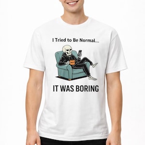 May include: White t-shirt with a graphic of a skeleton in a hoodie, seated on a blue armchair, holding a phone. The text "I Tried to Be Normal... IT WAS BORING" is printed above and below the graphic.