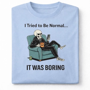 May include: Light blue t-shirt with the text "I Tried to Be Normal... It Was Boring." The graphic shows a skeleton in a hoodie sitting in a teal armchair, holding a phone and a coffee mug.