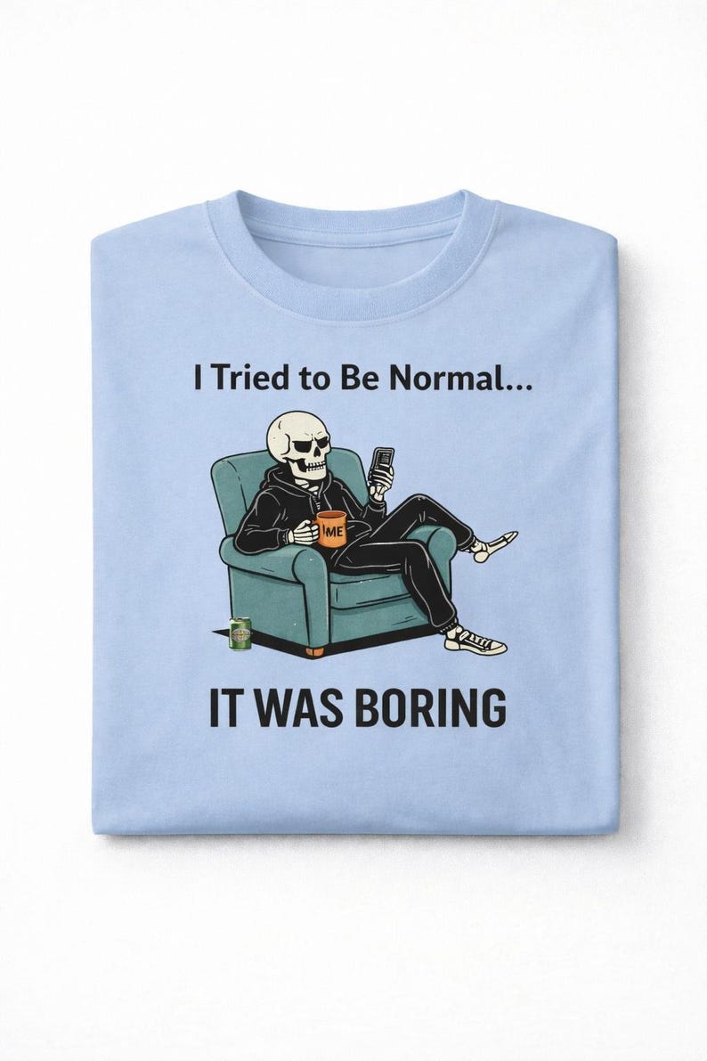 May include: Light blue t-shirt with the text "I Tried to Be Normal... It Was Boring." The graphic shows a skeleton in a hoodie sitting in a teal armchair, holding a phone and a coffee mug.