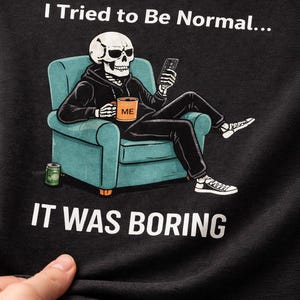May include: Black t-shirt featuring a skeleton in a hoodie, seated in a teal armchair, holding a phone and a mug that says "ME." The text "I Tried to Be Normal... IT WAS BORING" is printed above and below the illustration.