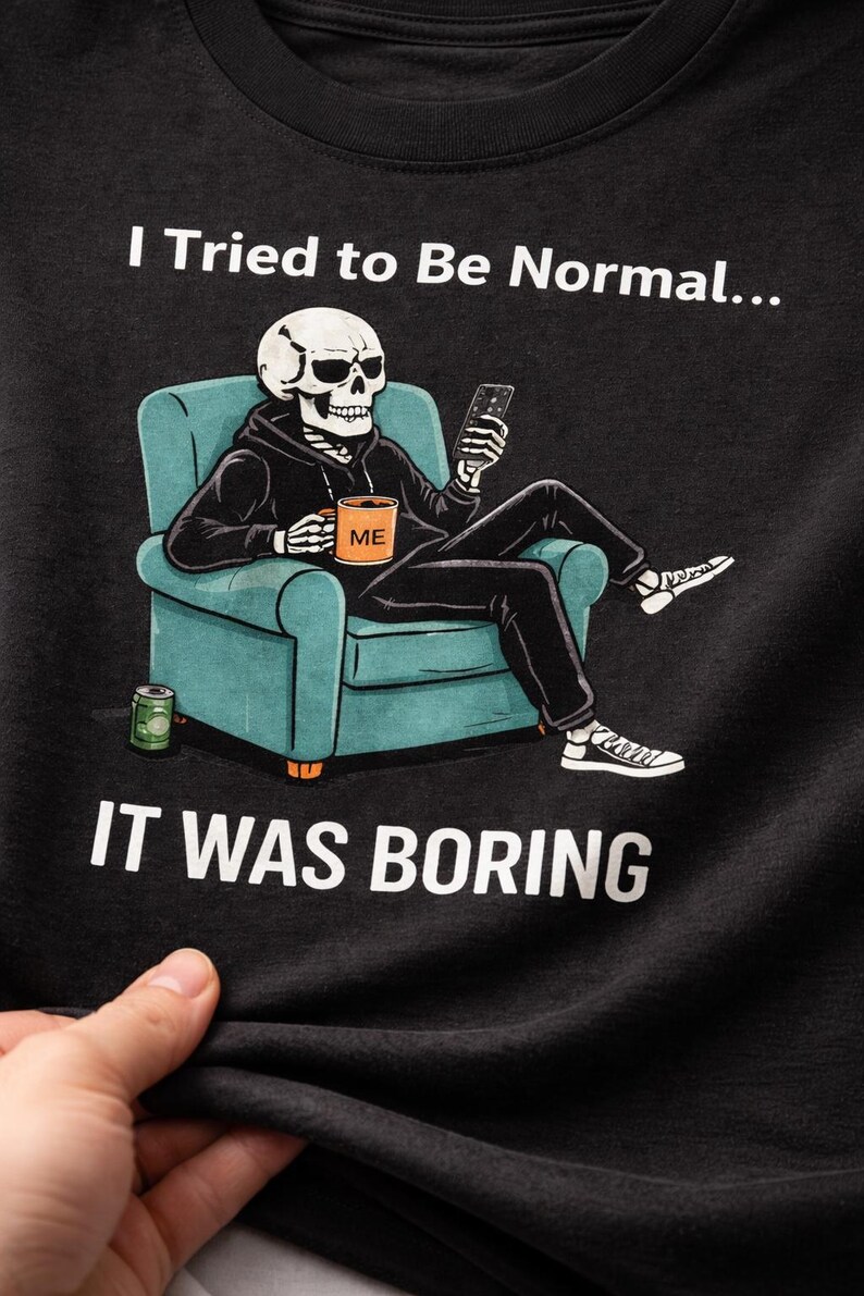 May include: Black t-shirt featuring a skeleton in a hoodie, seated in a teal armchair, holding a phone and a mug that says "ME." The text "I Tried to Be Normal... IT WAS BORING" is printed above and below the illustration.