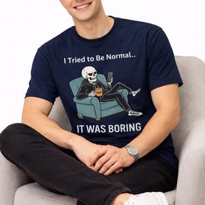 May include: Navy blue t-shirt with the text "I Tried to Be Normal.. IT WAS BORING" and a graphic of a skeleton relaxing on a chair. The skeleton is holding a phone and a mug. The person is wearing black pants and white sneakers.