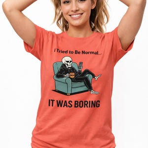 May include: Coral-colored t-shirt with the text "I Tried to Be Normal... IT WAS BORING." The shirt features a graphic of a skeleton in a hoodie, seated in a teal armchair, holding a phone and a coffee mug.
