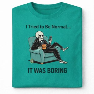 May include: Teal crew neck t-shirt with the text "I Tried to Be Normal... It Was Boring." The graphic shows a skeleton in a hoodie sitting in a chair, using a phone. The skeleton is holding a mug that says "TIME".