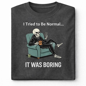 May include: Dark gray t-shirt with a humorous graphic. The design features a skeleton in a hoodie, seated in a teal armchair, holding a phone and a coffee mug that says "MEH." The text above reads "I Tried to Be Normal..." and below, "IT WAS BORING."