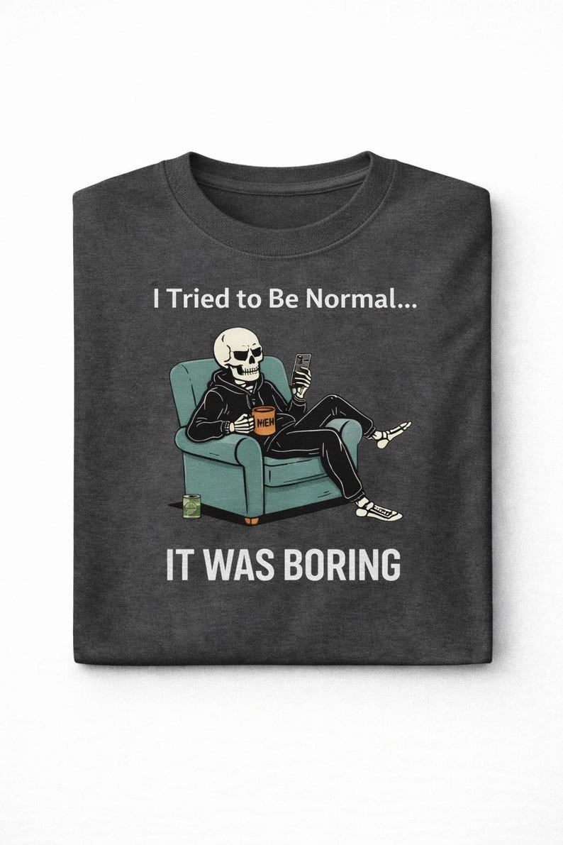 May include: Dark gray t-shirt with a humorous graphic. The design features a skeleton in a hoodie, seated in a teal armchair, holding a phone and a coffee mug that says "MEH." The text above reads "I Tried to Be Normal..." and below, "IT WAS BORING."