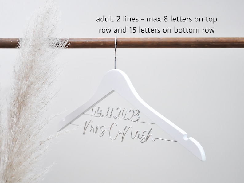 K&ouml;nnte beinhalten: Ein wei&szlig;er Holz Kleiderb&uuml;gel mit dem Text "04.11.2023" und "Mrs Chash" in Kursivschrift.