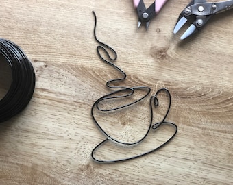 May include: A black wire is bent into the shape of a coffee cup with steam rising from the top. The wire is on a light brown wooden surface.