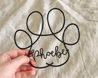 Może przedstawiać: Czarny druciany odcisk łapy z napisem "Phoebe" wewnątrz.