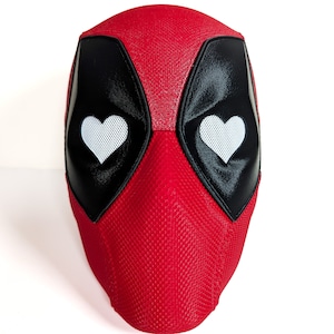 May include: A red and black Deadpool mask with white heart-shaped eye details. The mask has a textured, woven appearance and is designed to cover the entire face. The eye openings are black with white mesh and heart shapes.