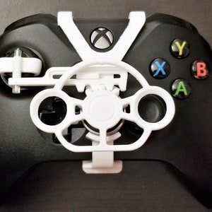May include: Black Xbox controller with a white steering wheel attachment. The controller has coloured buttons: green A, red B, blue X, and yellow Y. The white attachment has a circular design.