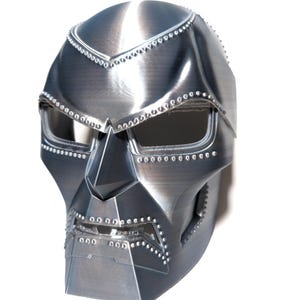 May include: A metallic silver mask with a distinctive angular design. The mask features eye openings, a prominent nose piece, and a series of rivets along the edges. The overall aesthetic suggests a futuristic or superhero theme.
