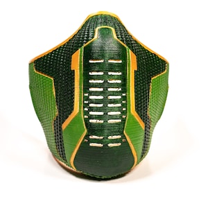 Op de afbeelding: Een groen en gouden gezichtsmasker met een geometrisch ontwerp. Het masker is gemaakt van een getextureerd materiaal en heeft een reeks kleine gaten aan de voorkant.