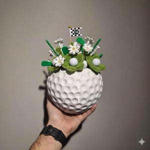 May include: A white golf ball-shaped vase filled with artificial flowers, golf balls, and tees. The arrangement includes green felt accents, white flowers, and a small flag with the number 18. The vase is held in a person's hand.