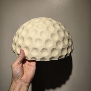 May include: A hemispherical object with a cream-colored surface featuring a repeating pattern of circular indentations, resembling the texture of a golf ball. The object has a smooth, matte finish and a scalloped bottom edge, held from below.
