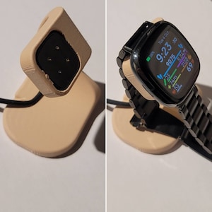May include: A beige 3D-printed smartwatch charging stand. The stand has a square charging cradle and a curved base. A black smartwatch with a black metal band is docked on the stand, displaying the time and fitness data. A black charging cable is attached.