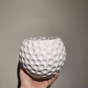 Op de afbeelding: Een witte, bolvormige decoratieve kom met een kuiltjesoppervlak, die lijkt op een golfbal. De kom wordt in een hand gehouden tegen een neutrale achtergrond. De open bovenkant van de kom suggereert dat deze gebruikt kan worden voor opslag of weergave.