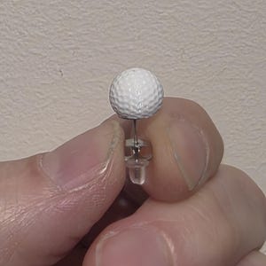 May include: A stud earring featuring a small, white, spherical golf ball charm with a textured dimpled surface. The charm is attached to a silver-toned metal post and includes a clear, translucent plastic earring back for secure fastening.