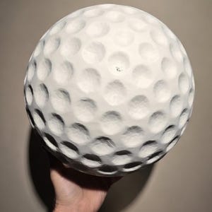 Könnte beinhalten: Ein großer, weißer Golfball mit strukturierter Oberfläche und Noppenmuster. Der Ball wird in einer Hand gehalten, um seine Größe und sein Design zu zeigen. Der Golfball ist ein klassischer Sportartikel.