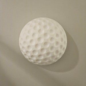May include: A white golf ball with a dimpled surface. The ball is round and appears to be made of a solid material. The golf ball is set against a neutral gray background.