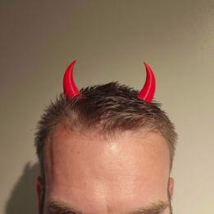 May include: A close-up of a person's head with short, dark hair. Two bright red devil horns are attached to the top of the head, curving upwards. The background is a plain, light-colored wall.
